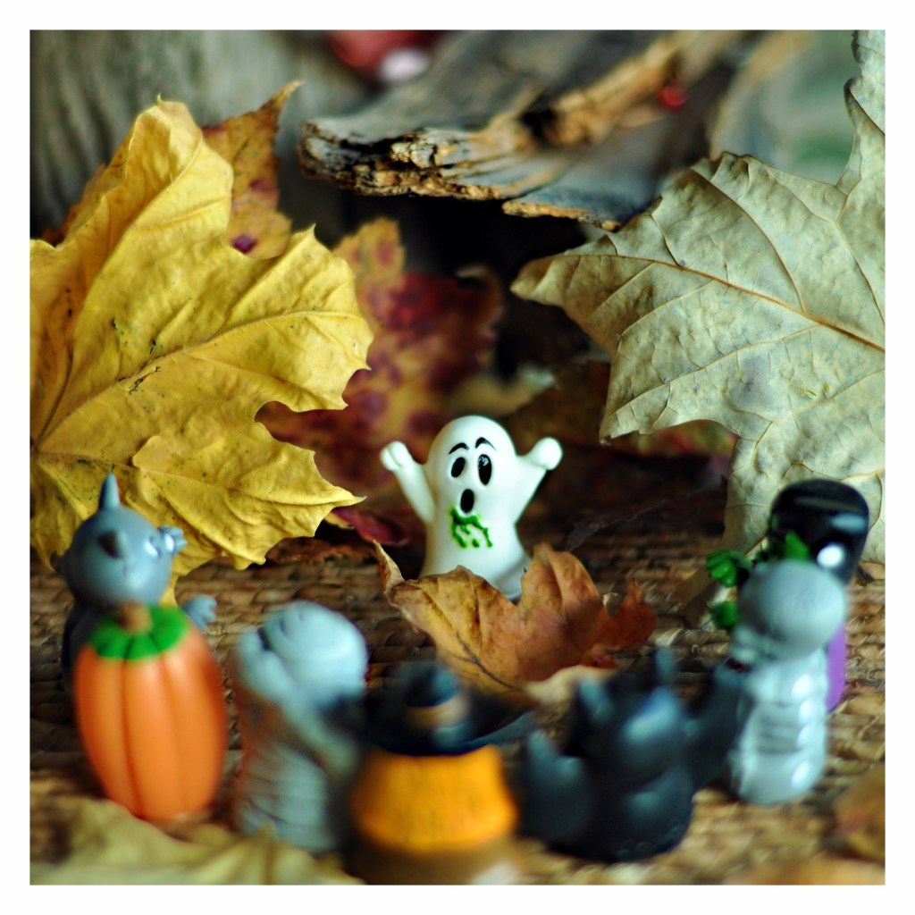 Tiny Halloween figures and a ghost arranged among fallen leaves and autumn decorations.